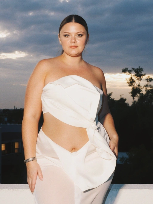 This is a confident young adult female model from the Americas, likely American, featuring dark brown hair pulled back neatly, fair skin, and a curvy body type. She exudes an elegant and self-assured vibe through her poised posture and stylish white asymmetrical outfit. The setting sun in the background complements her serene yet bold presence, making her highly suitable for fashion editorials, luxury brand campaigns, or lifestyle photography that celebrates elegance and confidence.